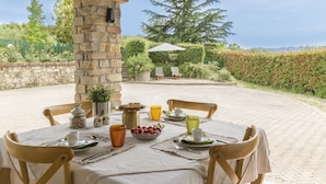Mesa redonda preparada para el desayuno, con un pilar de piedra, sillas exteriores y vistas al jardín y árboles.