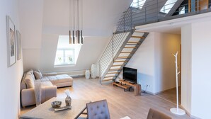 Een ruime woonkamer met een hoge plafond, een moderne trap, een flatscreen televisie en een eettafel met stoelen.
