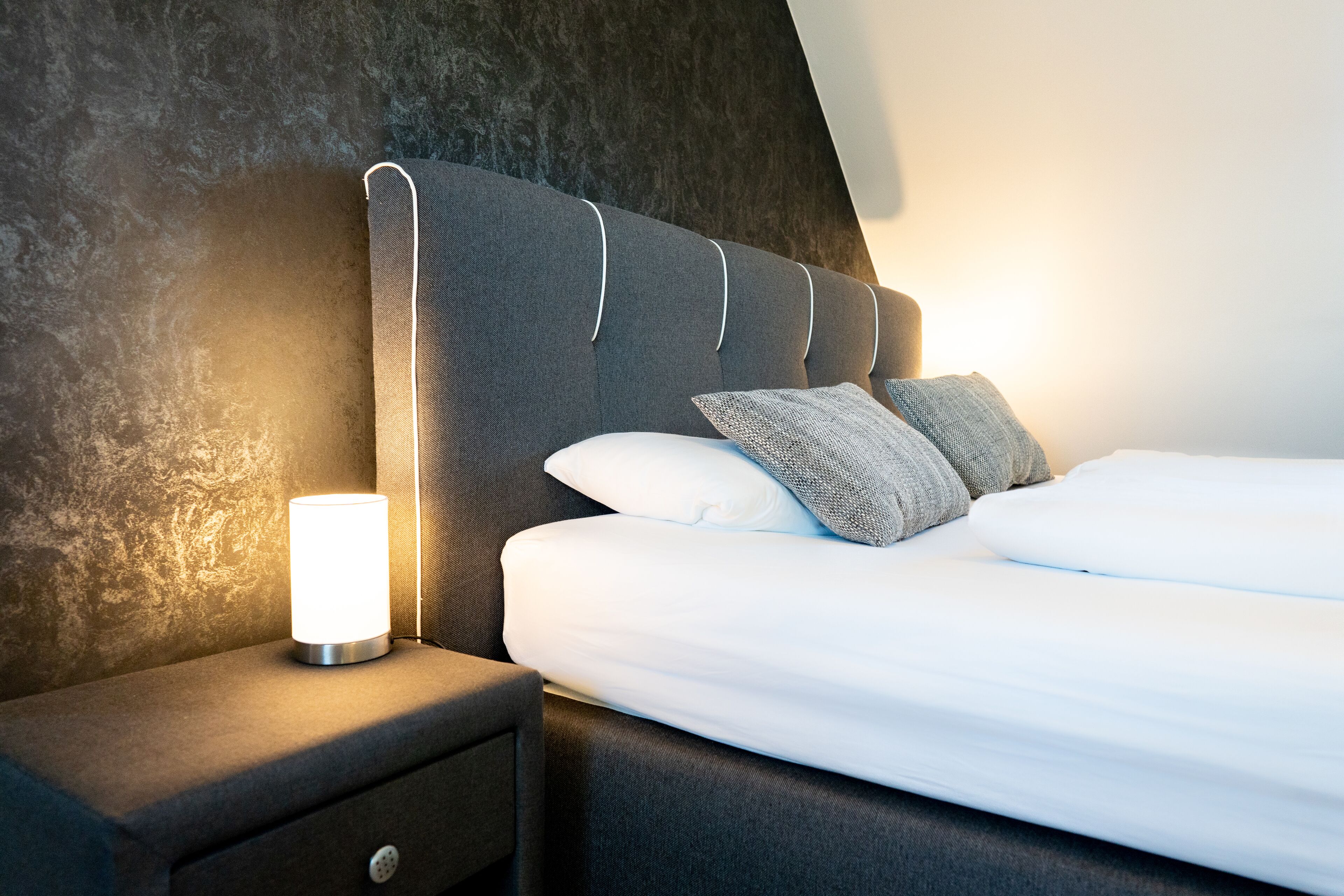 Een net opgemaakt bed met witte lakens en twee kussens, een bedlamp en een donkere, gestructureerde muur.