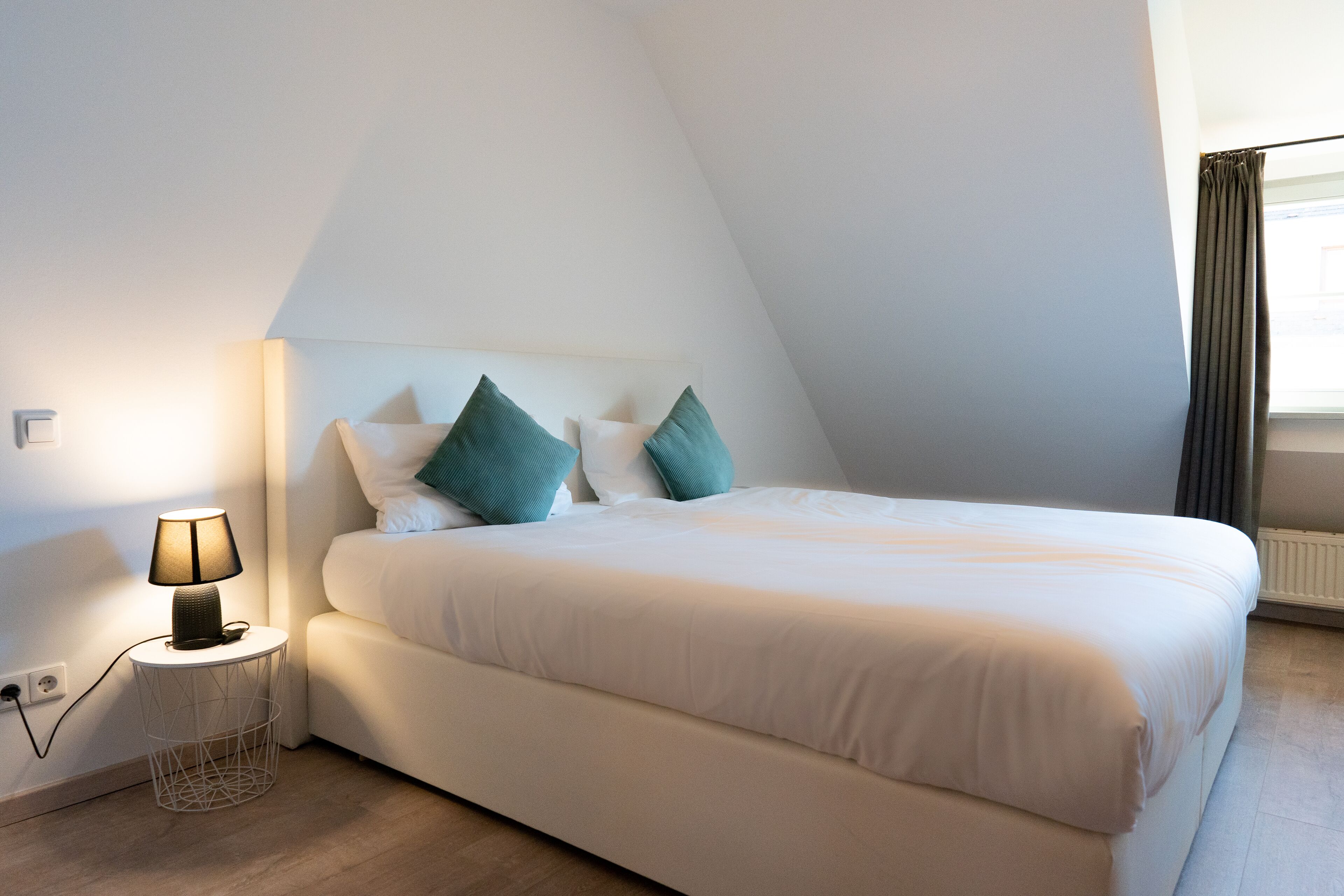 Ein ordentlich bezogenes Bett mit weißer Bettwäsche und türkisfarbenen Kissen, eine Nachttischlampe auf einem kleinen runden Tisch und ein Fenster mit Vorhängen.