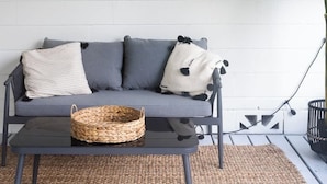 Un canapé gris, agrémenté d’un panier tressé et de deux coussins, est placé contre un mur orné d’un motif de feuilles de palmier et sur lequel on peut lire les mots « Je suis le plus heureux à la plage ».