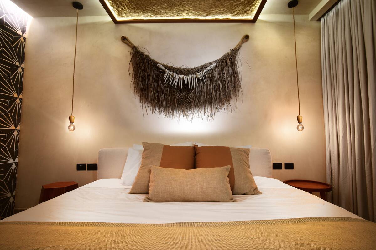 A modern hotel room with a large bed, two bedside tables, a hanging light fixture, and a unique wall decoration.