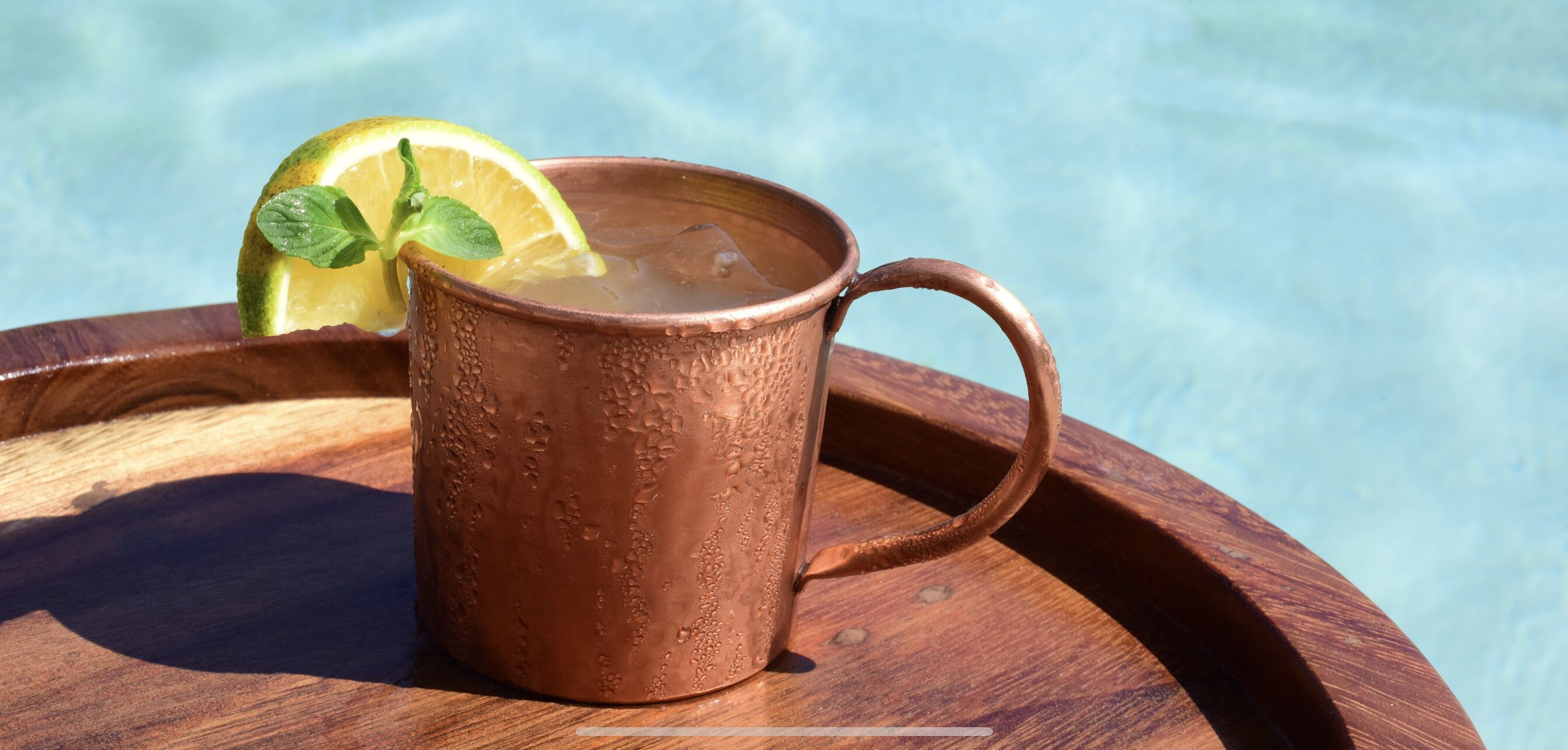 Un vaso de cobre con una rodaja de lima y menta sobre una bandeja de madera junto a una piscina.