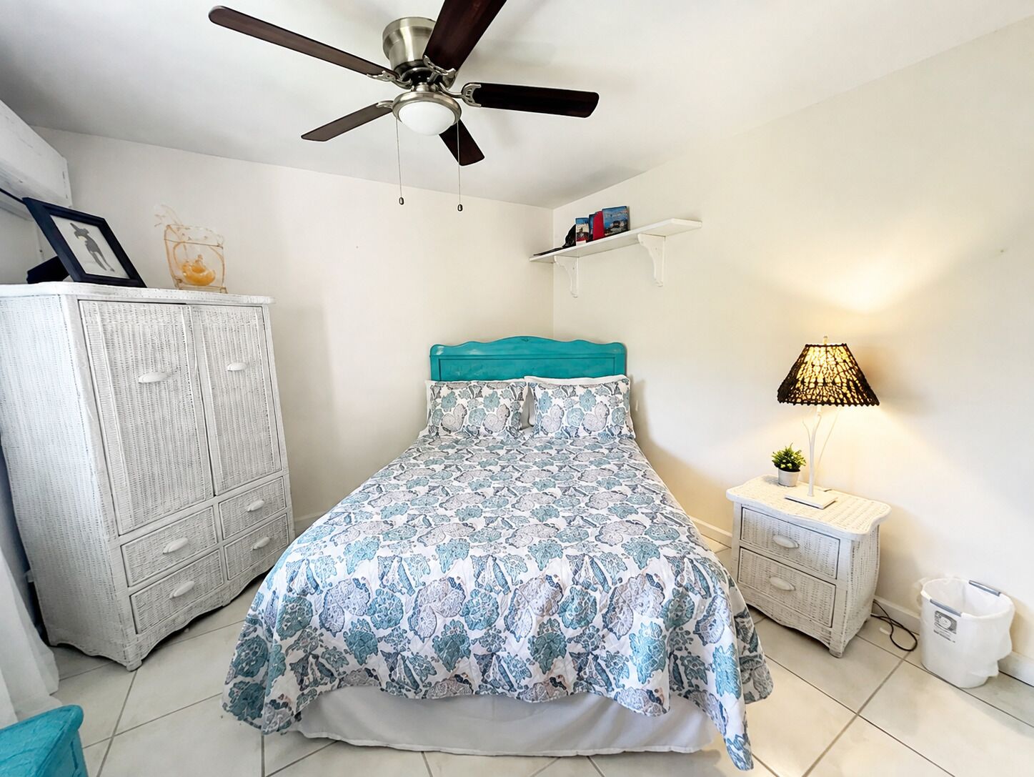 A bedroom with a bed, a ceiling fan, a nightstand with a lamp, a dresser, and a shelf with books.
