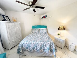 Ein Schlafzimmer mit einem Bett, einem Deckenventilator, einem Nachttisch mit Lampe, einer Kommode und einem Regal mit Büchern.