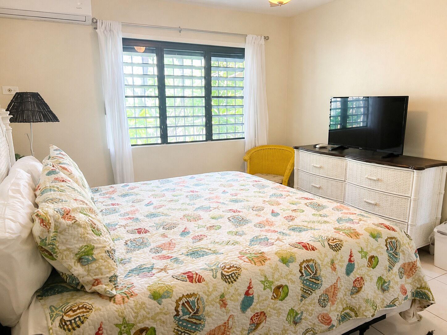 Ein Schlafzimmer mit einem Bett, einem Fernseher auf einer Kommode, einem Fenster mit Vorhängen und einem Sessel.