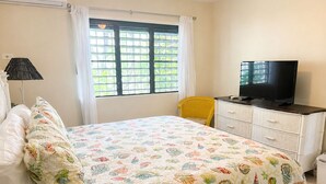 Un dormitorio con cama, televisor en una cómoda, ventana con cortinas y una silla.