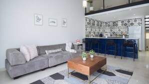 A modern living room with a grey sofa, a wooden coffee table, and a kitchen area with blue bar stools. - Beach Villages,  3 Bedrooms, Just Renovated (Humacao)
