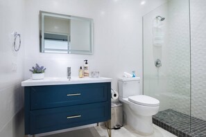 A modern bathroom with a white toilet, a blue vanity with drawers, a glass shower enclosure, and a small potted plant on the vanity. - Beach Villages, 3 Bedrooms, Just Renovated (Humacao)