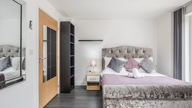 A bedroom with a large bed, a bedside table, a mirror, a wardrobe, and a shelf. - Beautiful 3-bed Apartment in Romford (Romford)