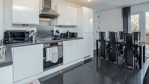 A modern kitchen with white cabinetry, a built-in oven, a microwave, and a coffee maker. There is a dining area with a table and chairs. - Beautiful 3-bed Apartment in Romford (Romford)