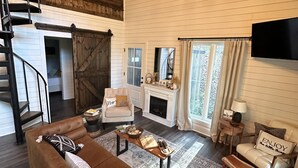 A spacious living room with a brown leather sofa, a fireplace, a television mounted on the wall, and a staircase leading to another level.