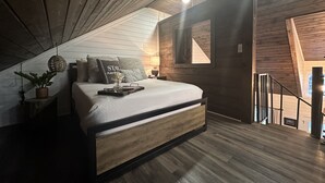 A bedroom with a sloped ceiling, wooden beams, a bed with pillows, a bedside table with a lamp, and a window with a view.