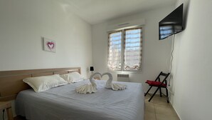 A neatly made bed with a heart-shaped pillow and two swan-shaped pillows, a TV mounted on the wall, and a chair with a red cushion. - App. One-bedroom apartment with all the amenities, pool, patio, Wi-Fi, 500 m from the beach, parking, air conditioning. (Santa-Maria-Poggio)