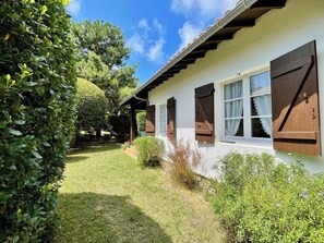 A single-story building with a white exterior and brown shutters, surrounded by well-maintained greenery and a clear sky. - Central Cap Ferret location, all on foot A0342 (Cap Ferret)