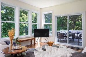 A living room with a large TV, a round coffee table, and a sofa. There are large windows and a sliding glass door leading to an outdoor deck. - 5BR Saugatuck-Area Home with Incredible Views (Douglas)