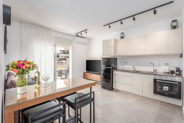 A modern kitchen with a glass dining table, black bar stools, a built-in oven, and a TV.