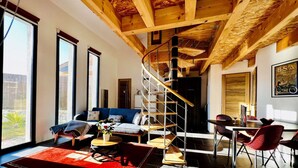 Un salón espacioso con techo de madera, un sofá azul, una mesa de comedor con sillas rojas y una escalera de caracol.