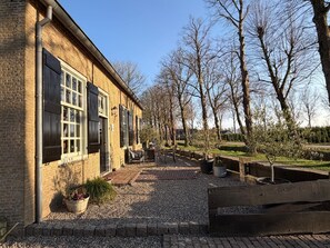 Ein Backsteinhaus mit großen Fenstern und Fensterläden, Außenbestuhlung und einem Kiesweg.