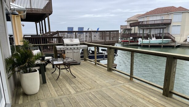 A wooden deck with a grill and outdoor furniture, overlooking a body of water and a building with a balcony. - North Padre Island waterfront canal home w/private pool and dock (Corpus Christi)