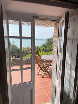 A view from a room with a sliding glass door to an outdoor patio with a table and chairs, overlooking a body of water and greenery. - Serra da Arrábida fantastic view (Setúbal)