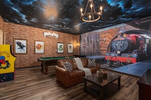 A room with a brick wall, a pool table, a leather sofa, and a large window with a mural of a train.