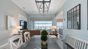 A modern living room with a glass dining table, white chairs, a brown sofa, a TV, and a large window with a view. - Extrended Pool Deck/No Rear Neighbors/Windsor Hills Resort, 3 Miles to Disney®! (Kissimmee)