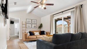 A spacious living room with a dark grey sofa, a beige sofa, and a wooden ladder against the wall. There is a ceiling fan, a large window with curtains, and a door leading to another room.