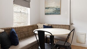 A room with a round table, two chairs, a wicker sofa, a window with a view of a brick wall, and a painting on the wall.