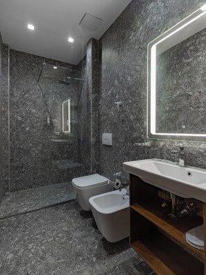Un baño moderno con pared de mármol, ducha con cerramiento de vidrio, lavamanos blanco y un inodoro.