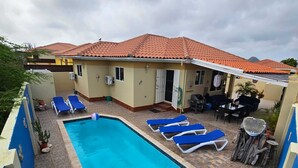 Una piscina con sillas reclinables azules, un patio techado y un edificio con tejado de tejas rojas.
