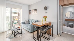 A modern living room with a sofa, a dining table with chairs, a coffee table with fruit, and a view of the city through large windows. - Superb 1Bdr w balcony (Jerusalem)
