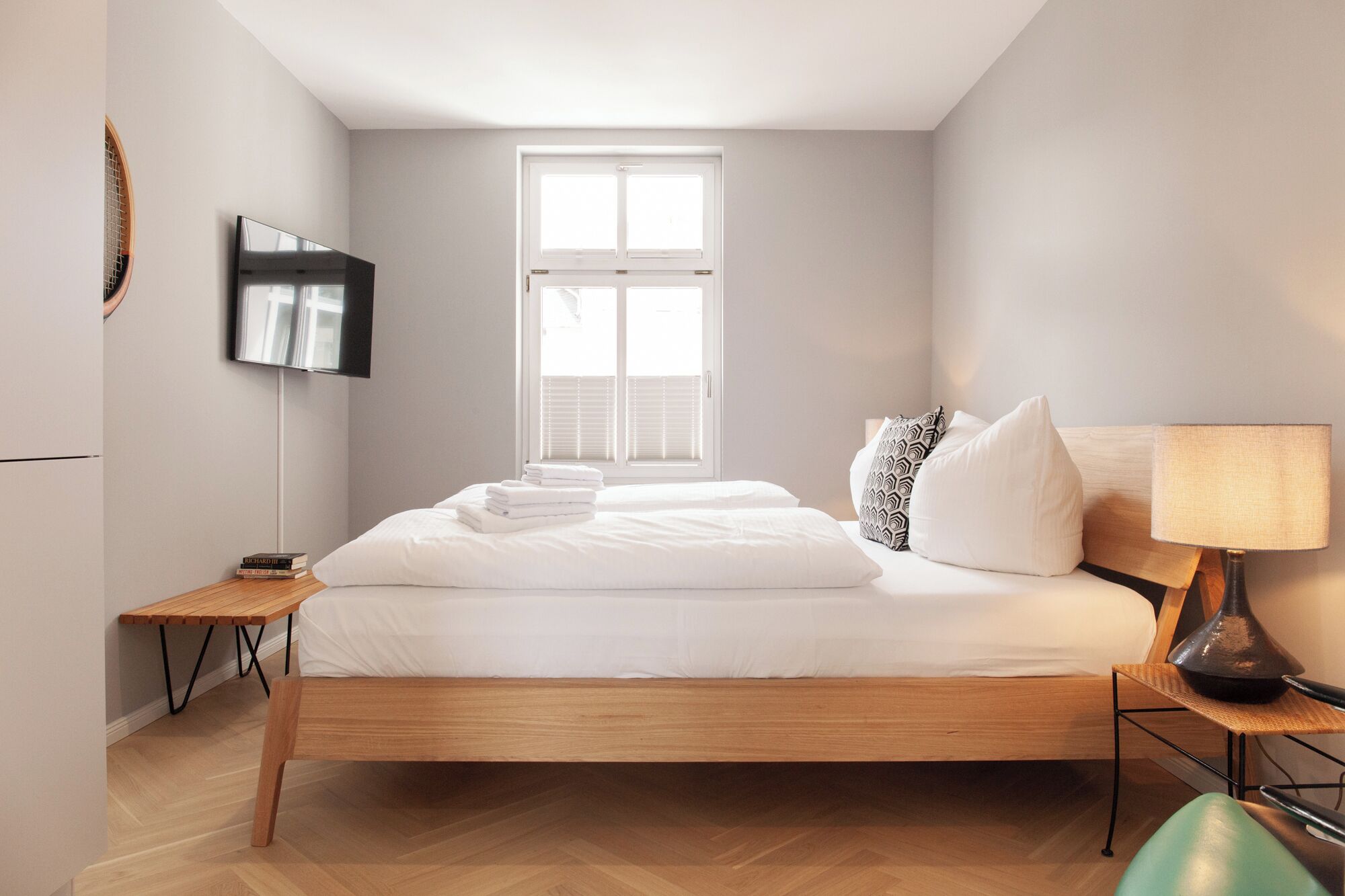 Un dormitorio moderno con una cama de madera, un televisor de pantalla plana montado en la pared, una ventana con persianas y una mesita de noche con lámpara.