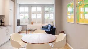 Een modern appartement met keuken, eethoek en woonkamer. Het heeft grote ramen, een ronde eettafel en een bank.