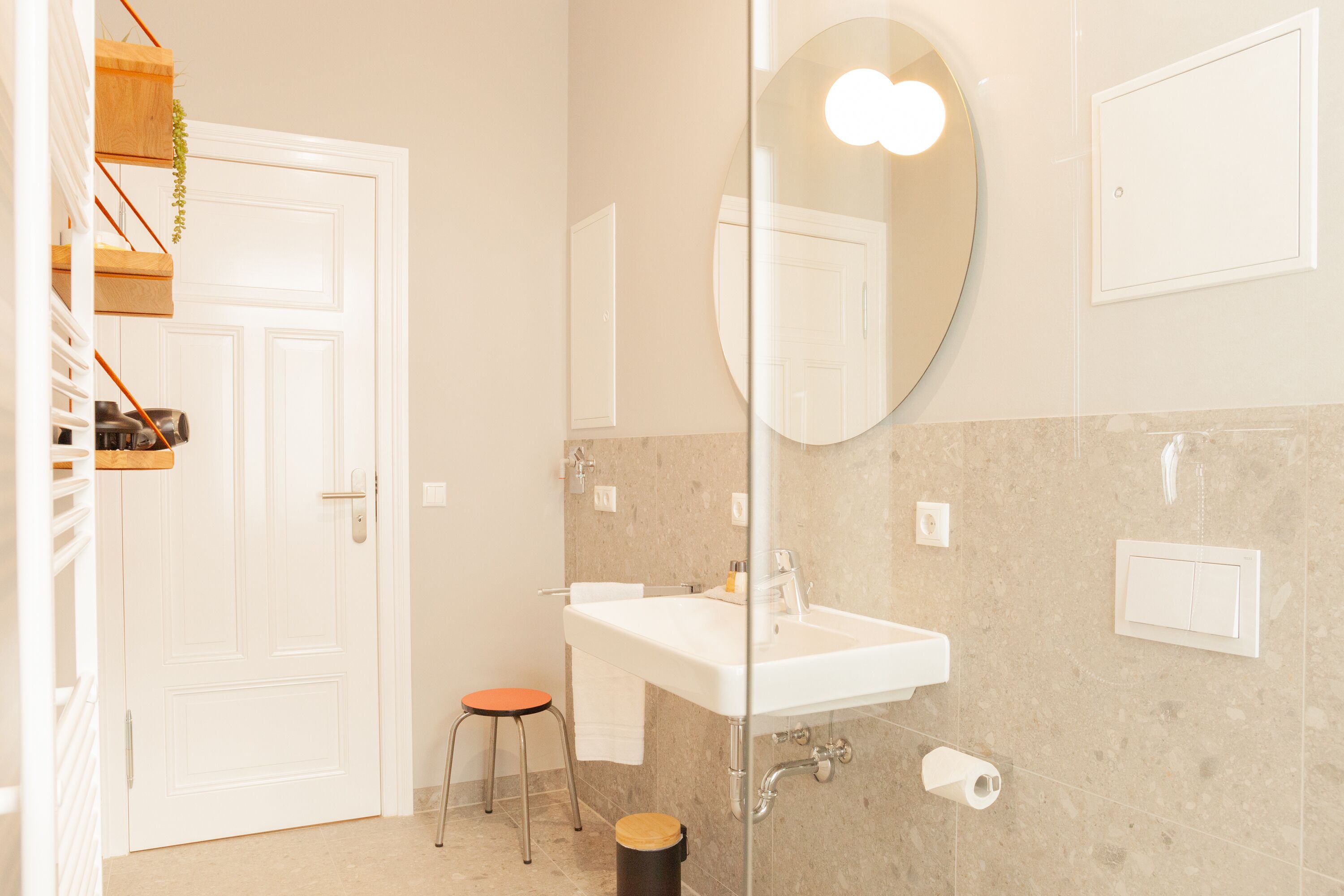 Ein Badezimmer mit einem weißen Waschbecken, einem Spiegel und einer Toilette. Ein Holzregal mit einer Pflanze und einem Hocker sind ebenfalls vorhanden.