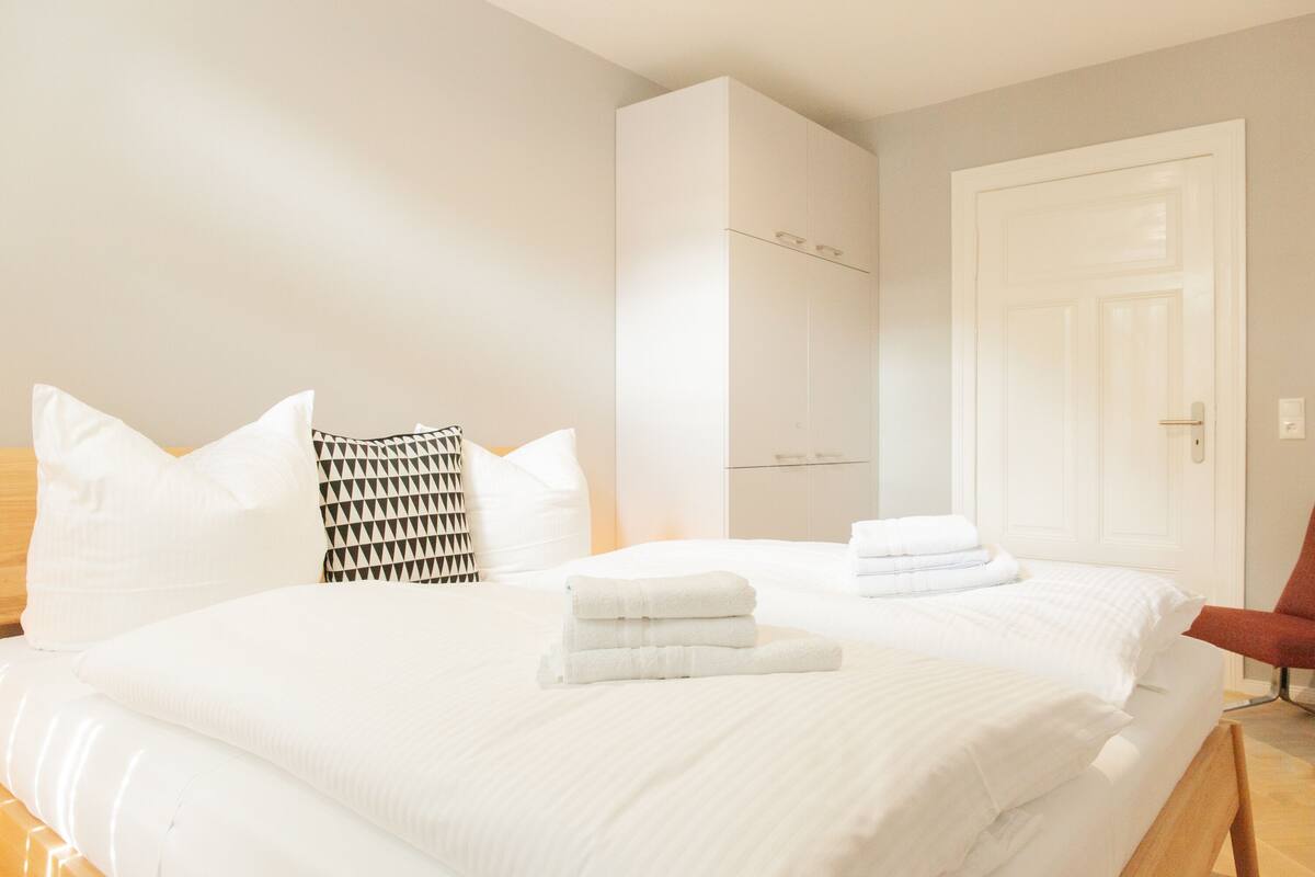 Habitación de hotel con una cama grande, ropa de cama blanca, cabecero de madera, una silla roja y un armario blanco.
