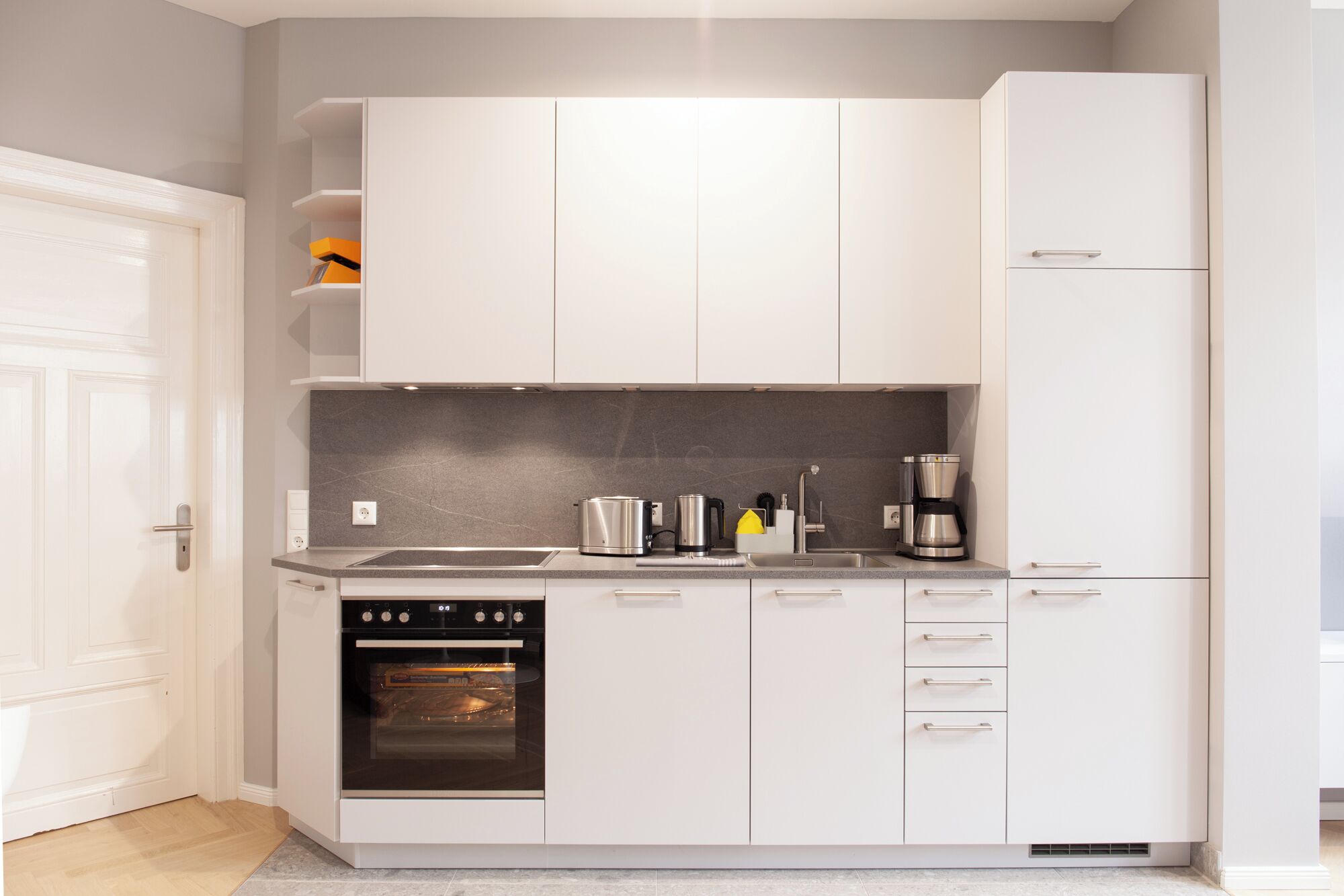 Una cucina compatta con mobili bianchi, forno integrato, piano cottura e macchina da caffè.