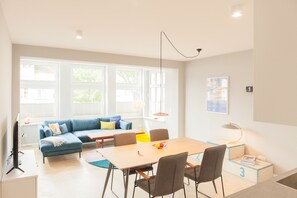 Ein modernes Wohnzimmer mit einer Couch, einem Esstisch und Stühlen. Es gibt große Fenster, eine Lampe und ein Gemälde an der Wand.