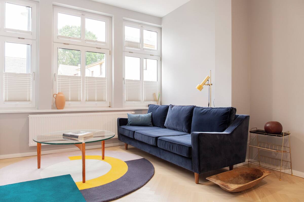 Una sala de estar moderna con un sofá azul, una mesa de centro de vidrio, una lámpara amarilla y un cuenco de madera en el suelo.