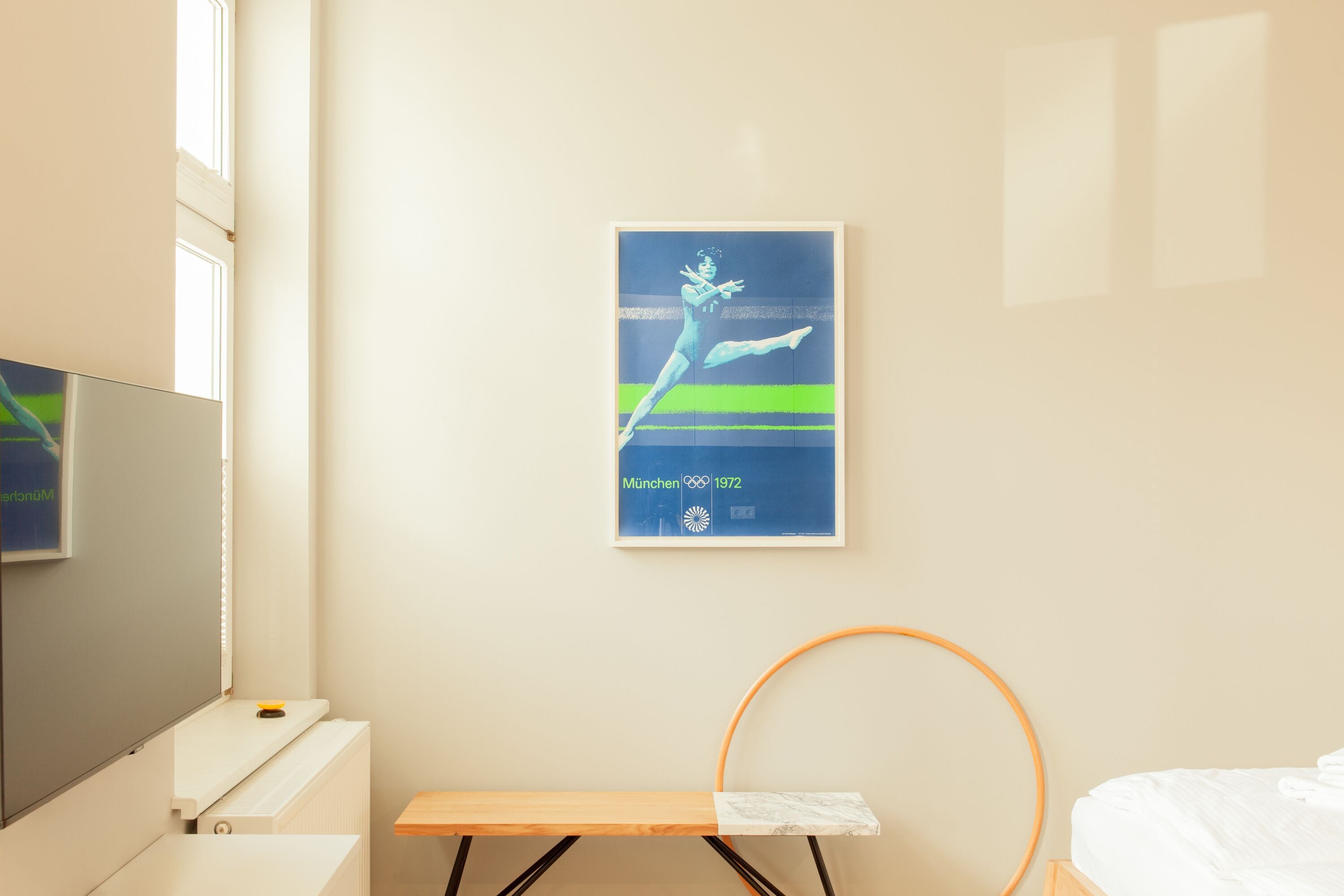 A hotel room with a bed, a small table, a wall-mounted TV, and a poster on the wall.