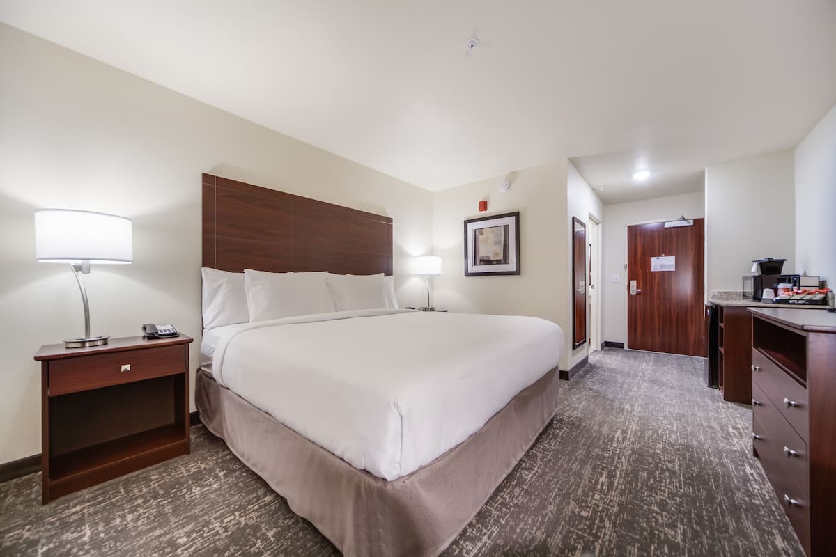 A hotel room with a large bed, bedside tables with lamps, a wooden headboard, a framed picture on the wall, a desk with a computer, and a door leading to another room.