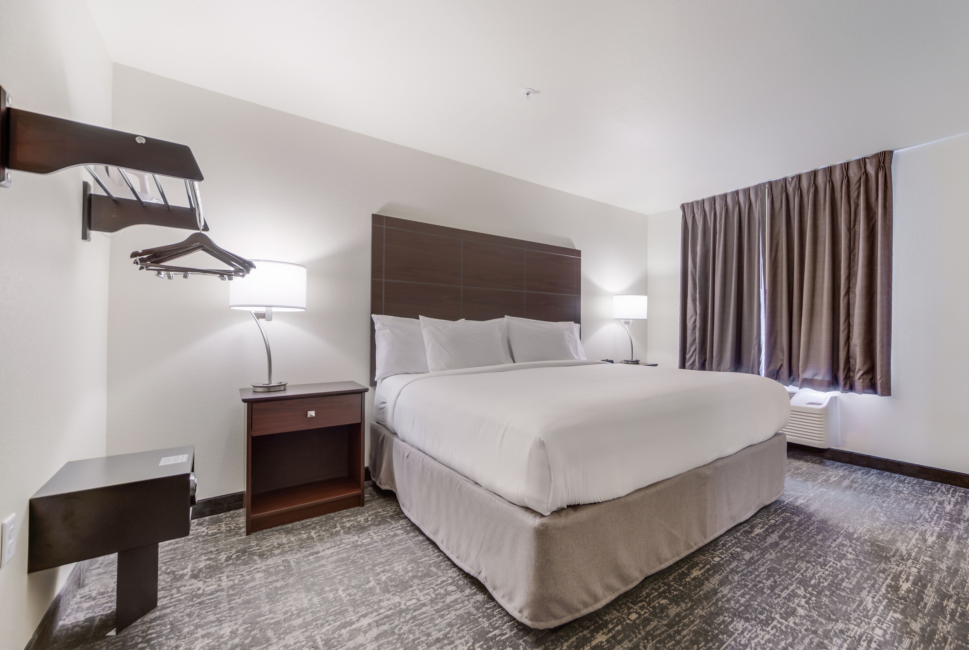 A hotel room with a large bed, two bedside tables, a wall-mounted coat rack, and a window with curtains.