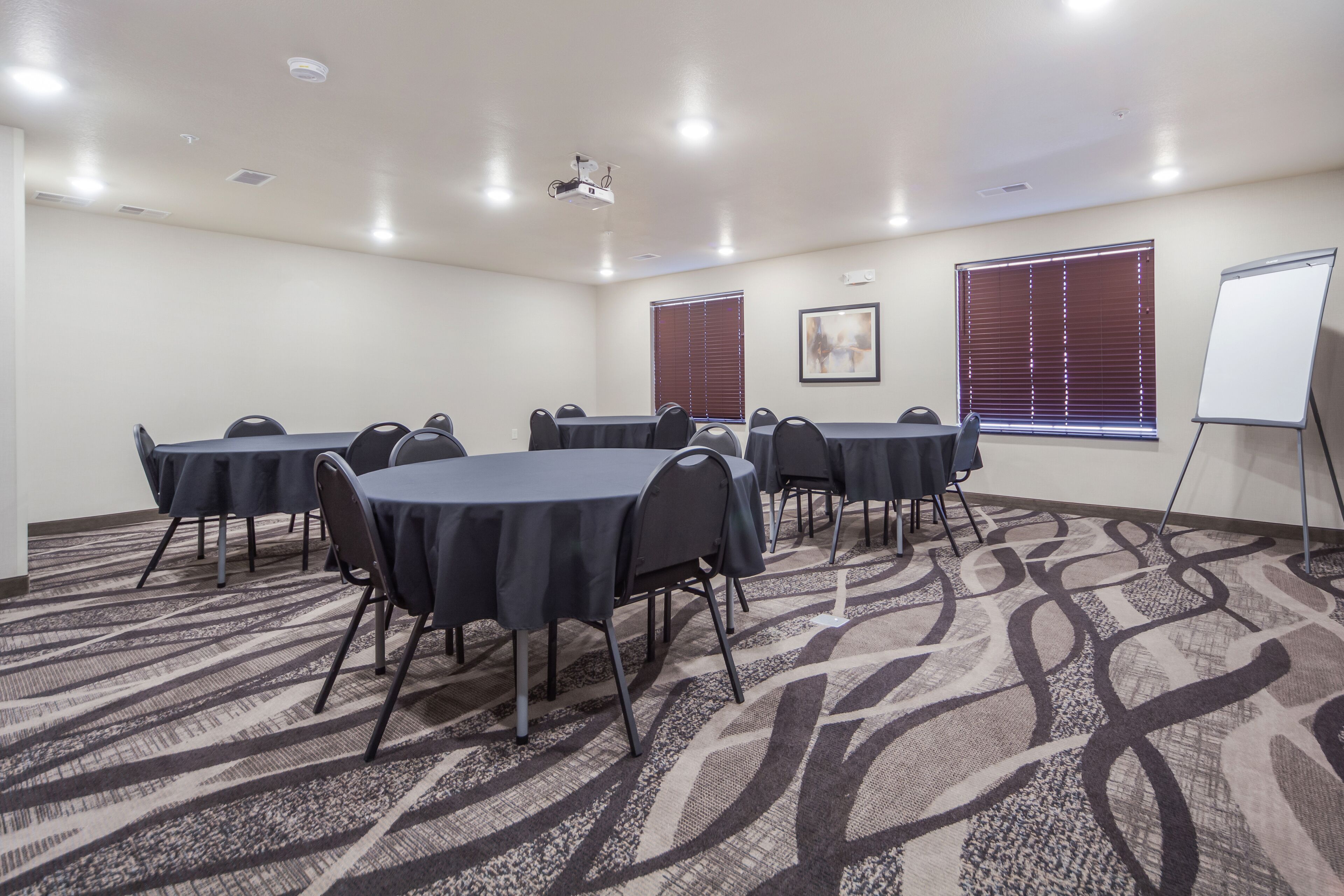 Un salón de conferencias con suelo alfombrado, mesas con sillas, un proyector, una pizarra y ventanas con persianas.