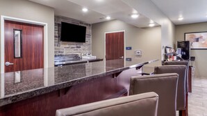 A modern kitchen with a granite countertop, a stone backsplash, and a flat-screen TV mounted above the island. There are two wooden doors and a refrigerator in the background. - Cobblestone Inn & Suites-Fremont (Fremont)