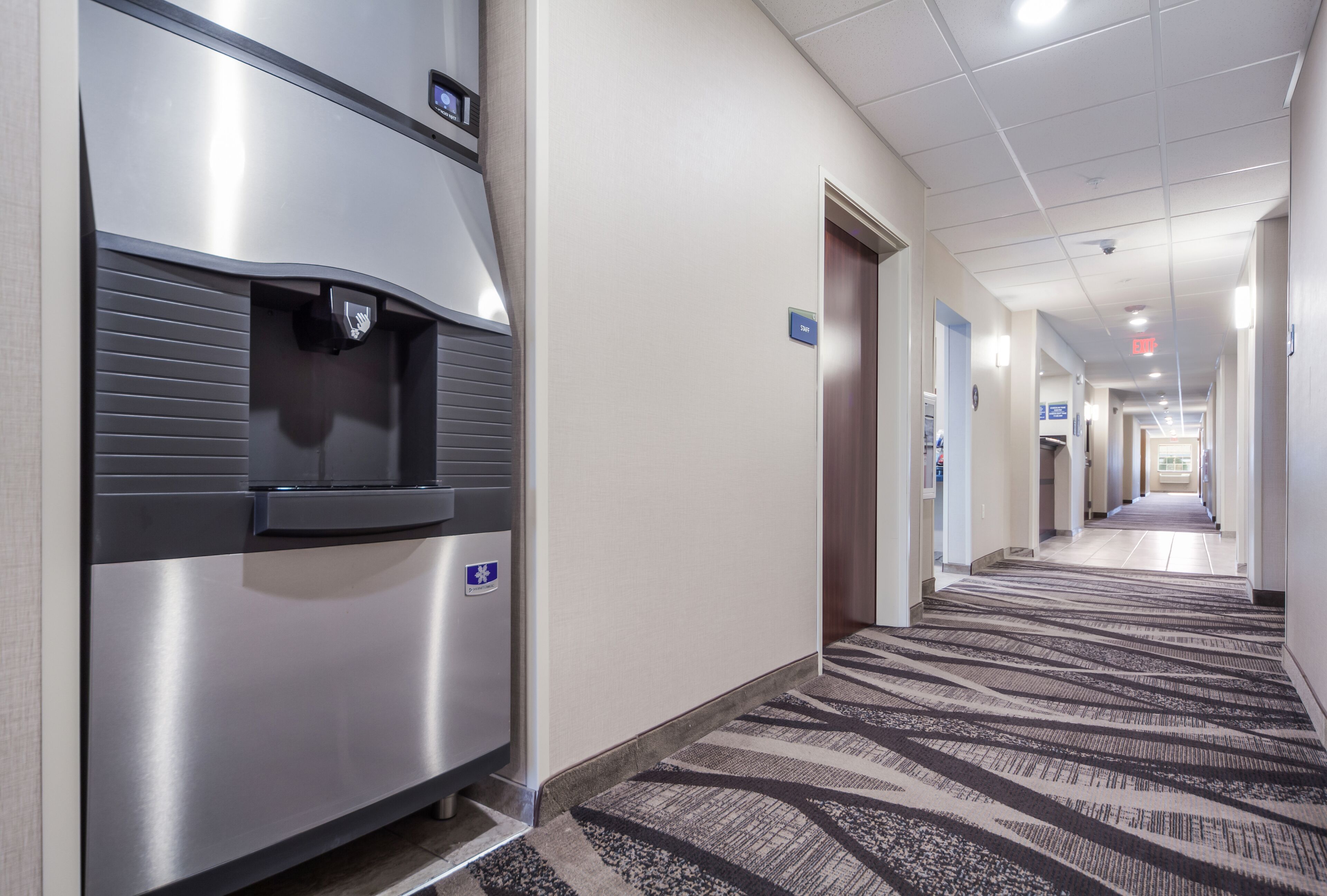 A hotel corridor with a water dispenser, a carpeted floor, and a sign indicating the direction to the elevator.