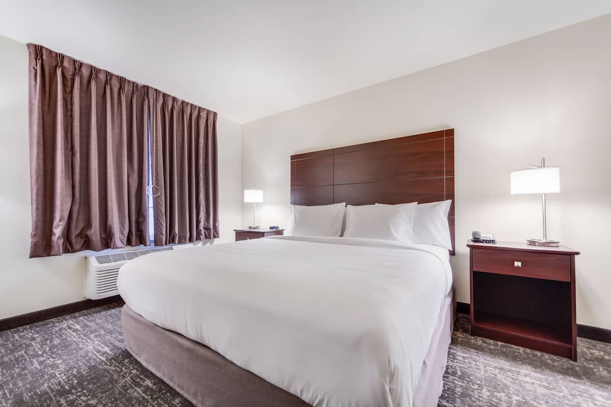 A hotel room with a large bed, two bedside tables with lamps, a window with curtains, and a wall-mounted heater.