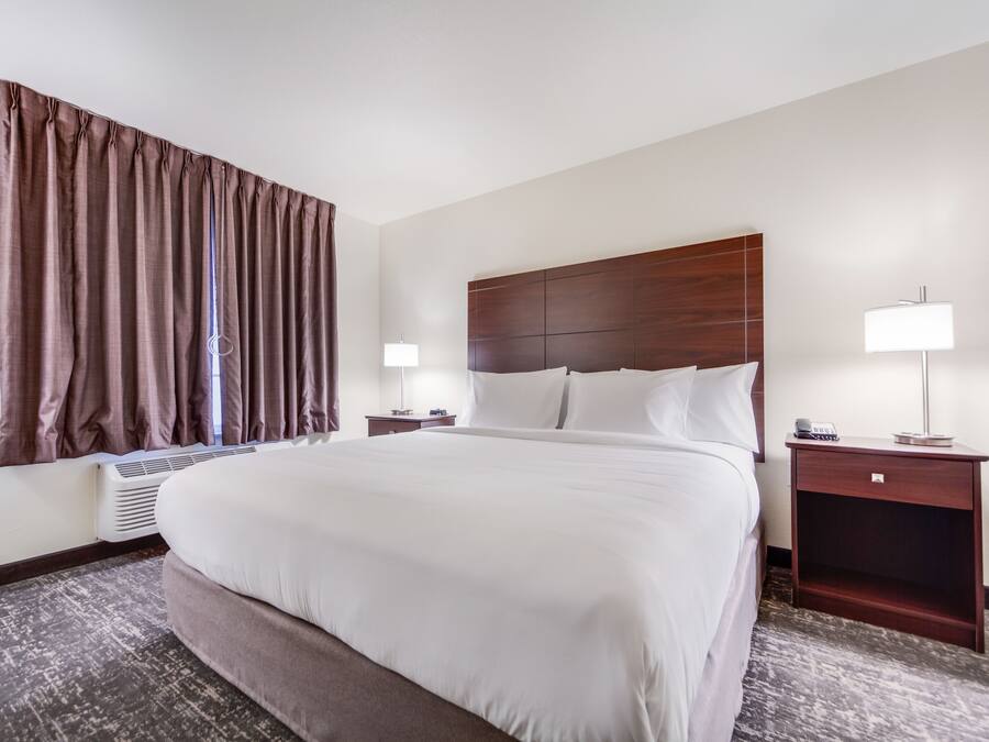 A hotel room with a large bed, two bedside tables with lamps, a window with curtains, and a wall-mounted heater.