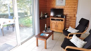 Una sala de estar acogedora con detalles de madera, un televisor sobre un mueble de madera, una zona de comedor con mesa y sillas, y un ventanal corredero que da a un patio exterior.