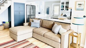 Ein modernes Wohnzimmer mit einer Couch, einem kleinen Holztisch, einem Streifen Teppich und einer Küchenzeile mit Kühlschrank und Schränken.