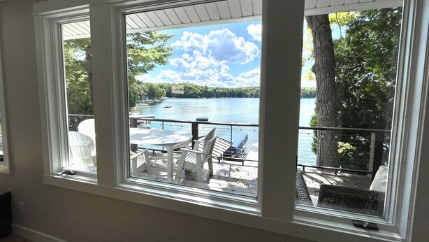 Ein Zimmer mit großem Fenster und Seeblick, eine Terrasse mit Gartenmöbeln sowie Blick auf Boote und Bäume.